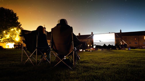 Two visitors sitting in folding chairs at dusk with a film showing on an outdoor screen at a cinema experience at Hardwick, Derbyshire.